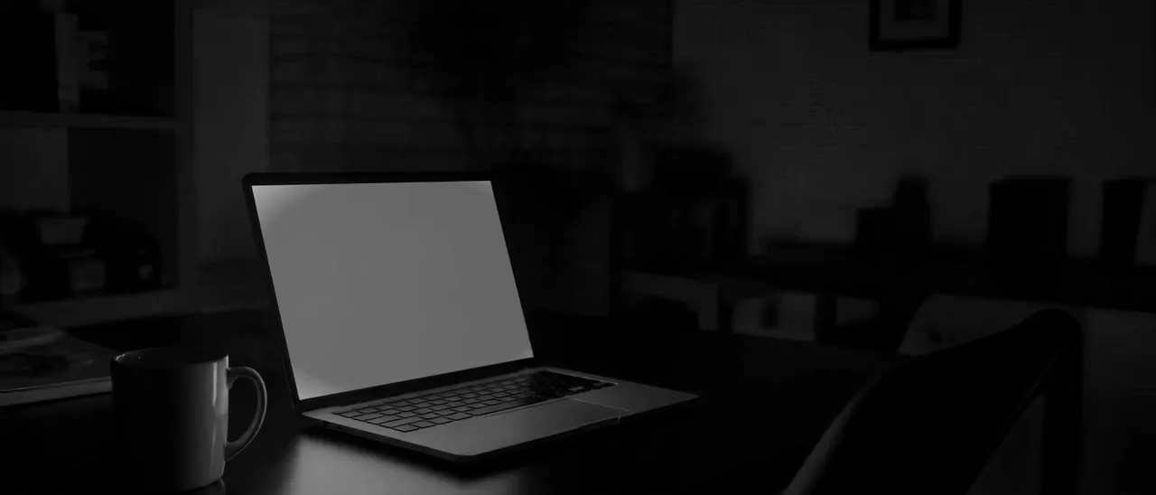 A laptop open on a dark table at night, its screen the only light in the room, a mug beside it.